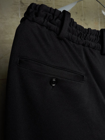 Close-up of black trousers featuring an elasticized waistband and a buttoned welt pocket, hanging against a neutral textured background.