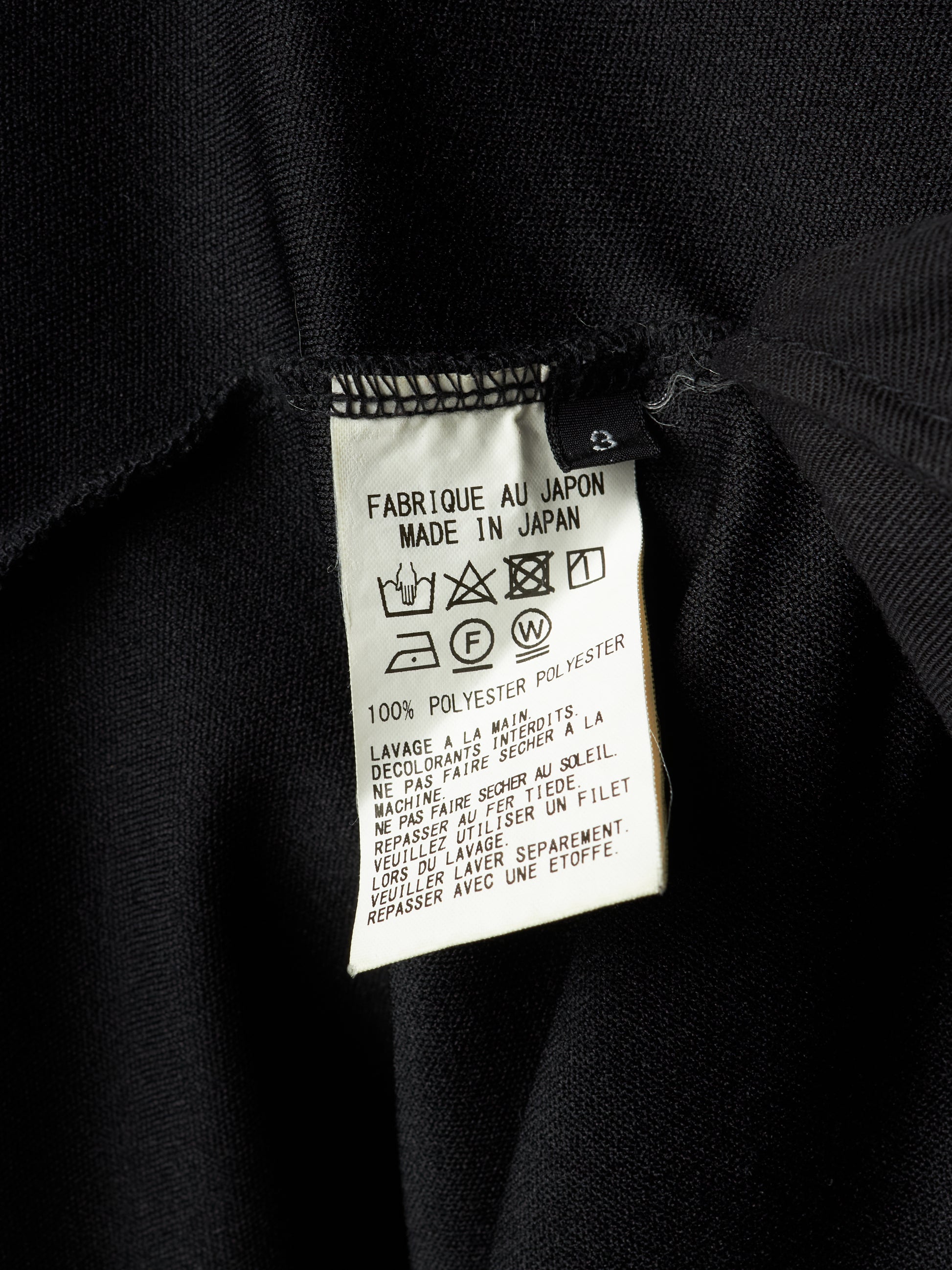 Close-up of a black garment's interior label showing 'FABRIQUE AU JAPON MADE IN JAPAN', '100% POLYESTER', and care symbols on a white paper tag.