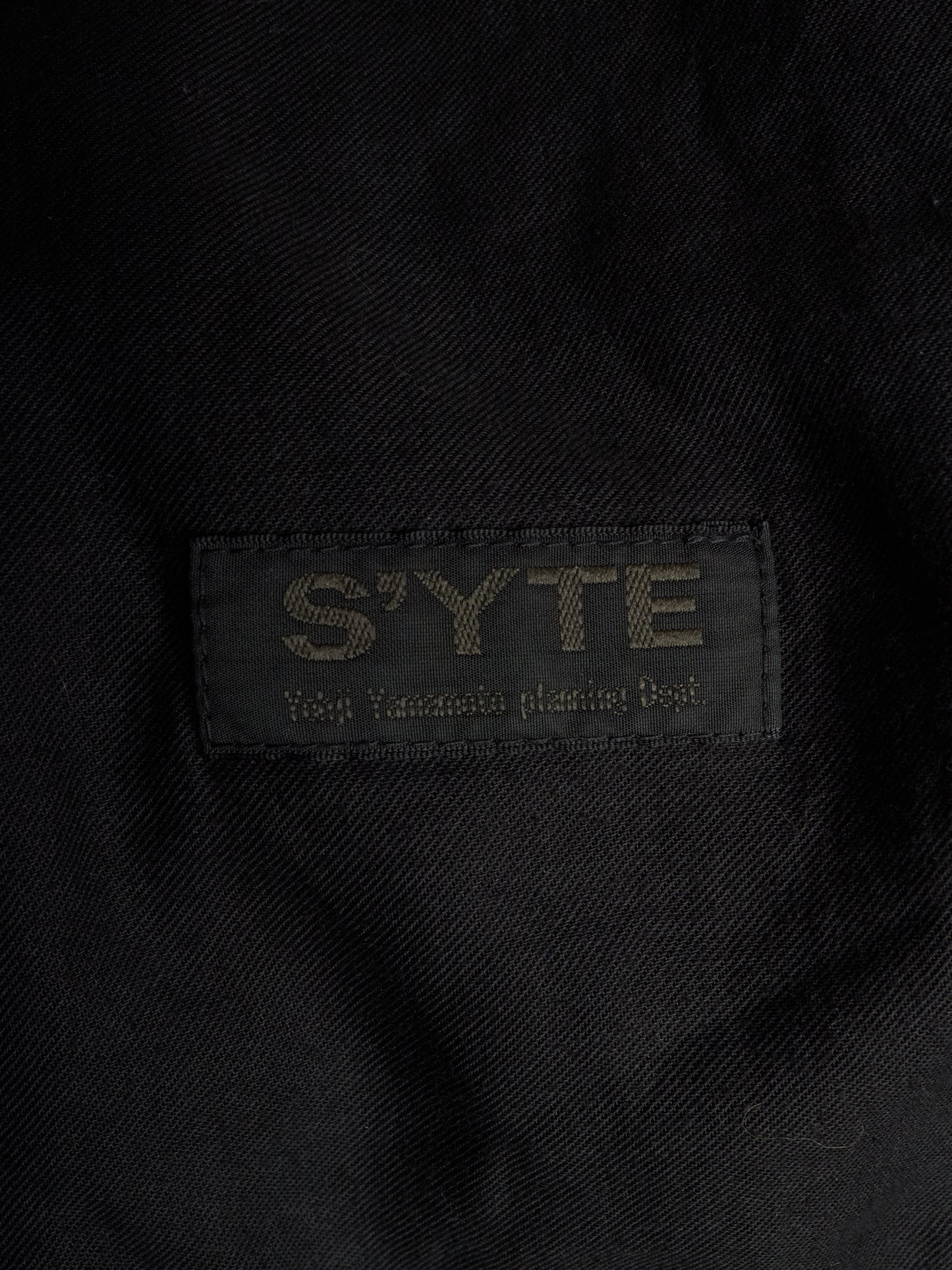 Close-up of a black woven fabric label with the brand name 'S'YTE' and the text 'Yokohama plastic bag' stitched in a contrasting light tone.