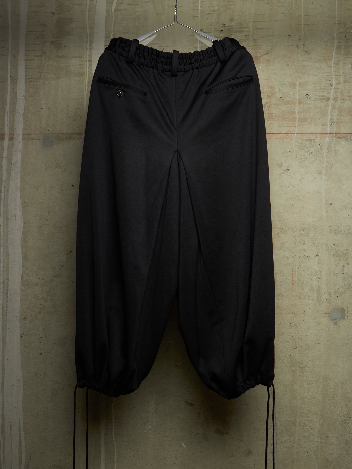 Black wide-leg jogger pants with an elastic waistband and drawstring hems, hanging against a textured concrete wall.