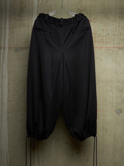Black wide-leg jogger pants with an elastic waistband and drawstring hems, hanging against a textured concrete wall.