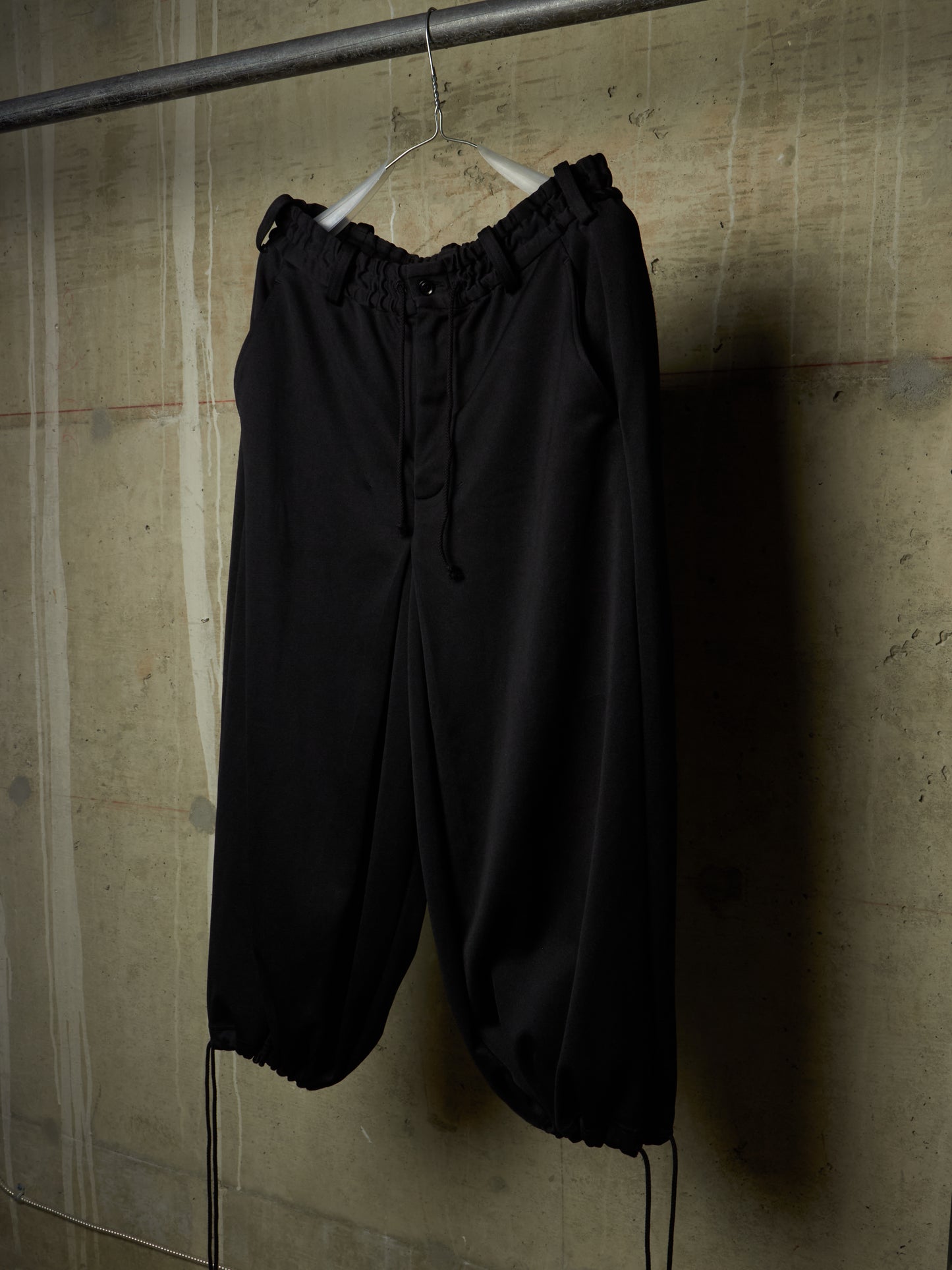 Black wide-leg cropped pants with an elastic drawstring waist and gathered cuffs, hanging on a white hanger against a textured concrete wall.