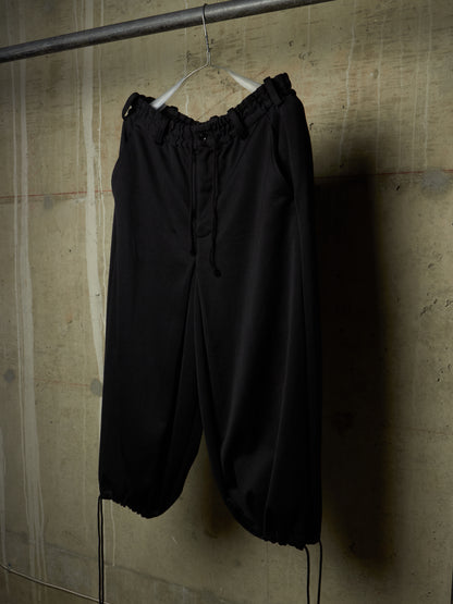 Black wide-leg cropped pants with an elastic drawstring waist and gathered cuffs, hanging on a white hanger against a textured concrete wall.