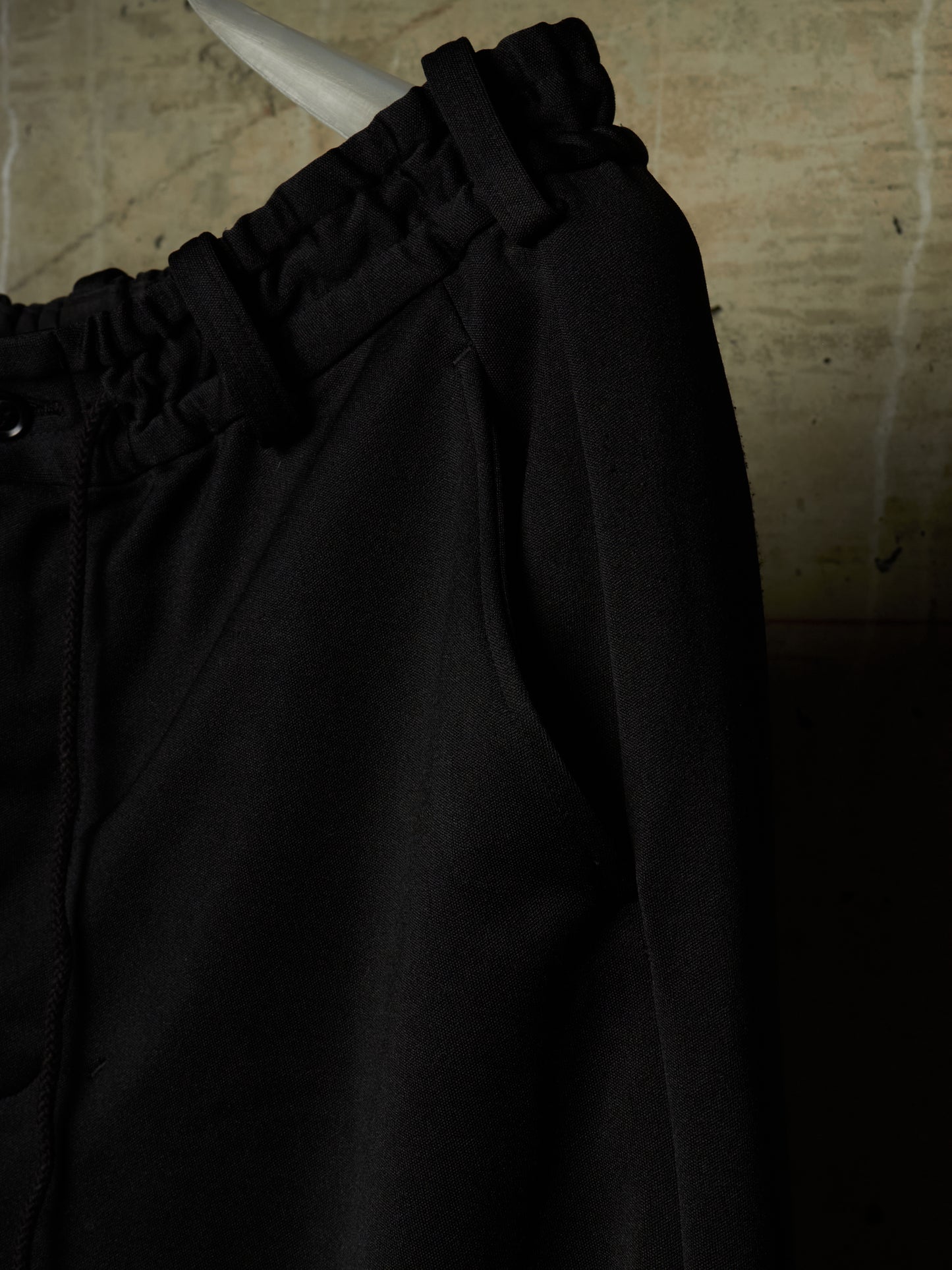 Close-up of black drawstring pants hanging against a textured beige wall, showing the elastic waistband and fabric texture.