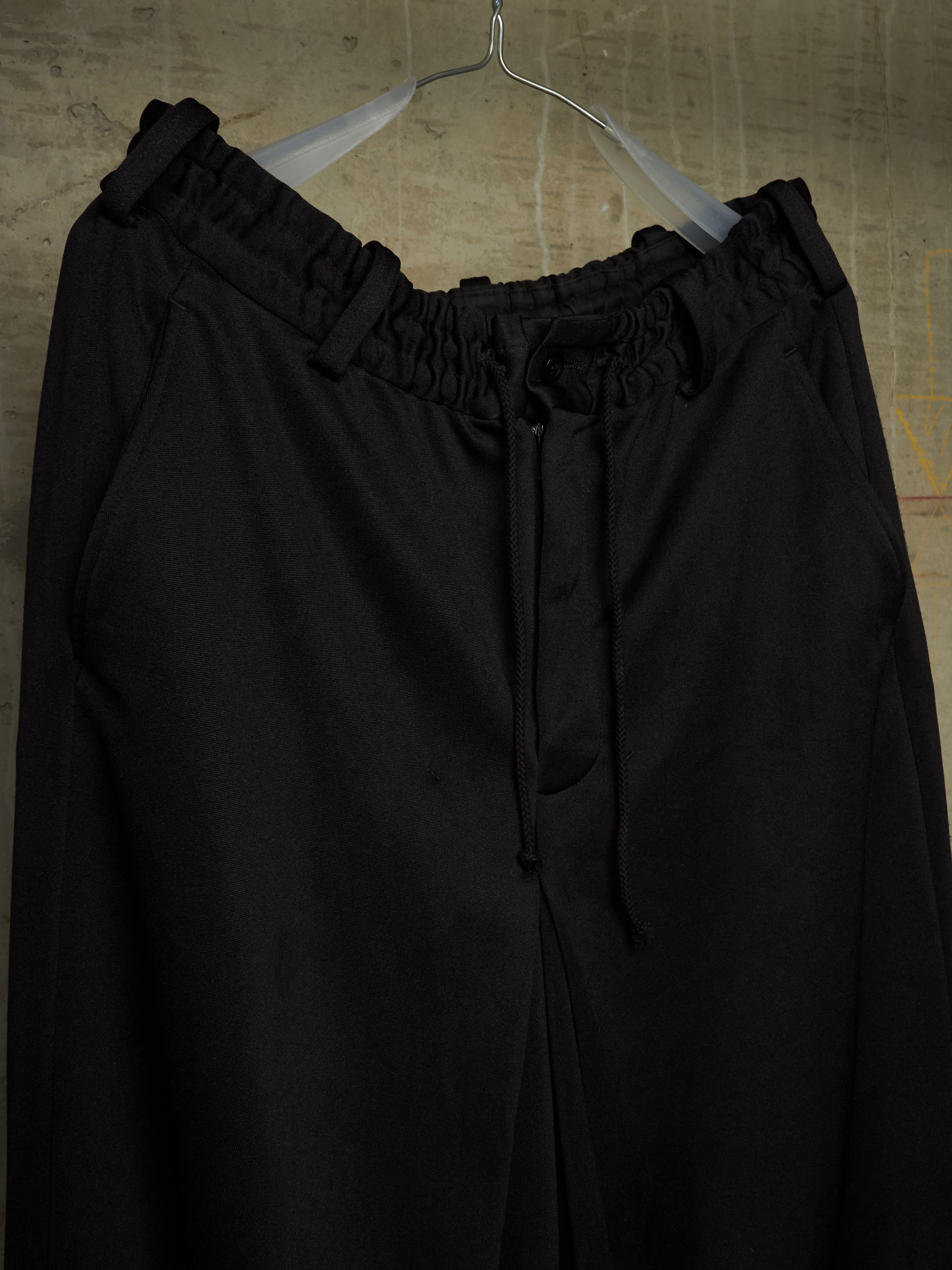 Black wide-leg drawstring pants with an elasticized waistband, hanging on a clear plastic hanger against a textured beige wall.