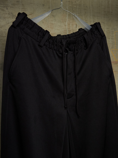 Black wide-leg drawstring pants with an elasticized waistband, hanging on a clear plastic hanger against a textured beige wall.