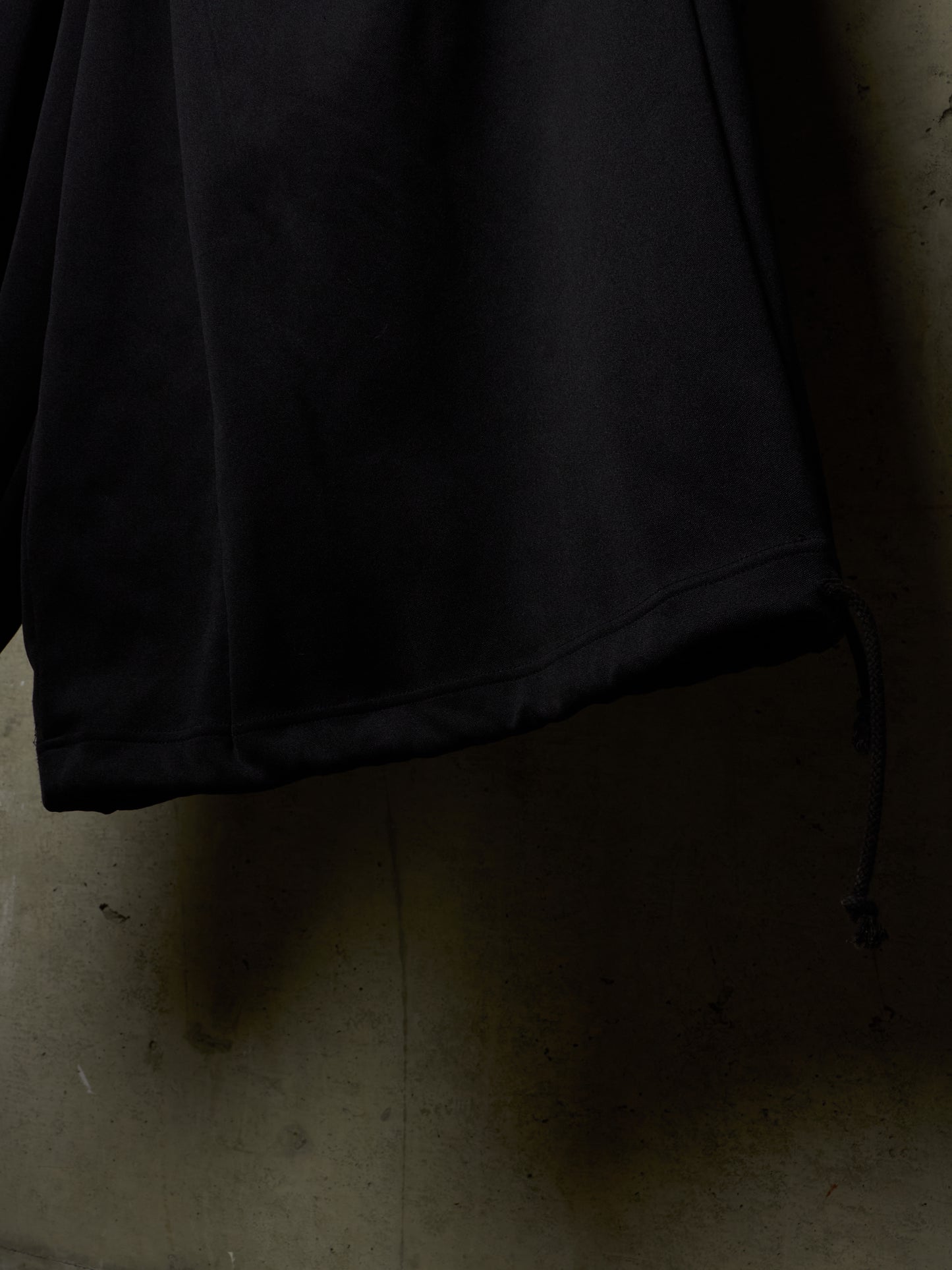 Close-up of a black fabric garment, likely a jacket or coat sleeve, hanging against a dimly lit concrete wall with a drawcord detail visible near the bottom right.