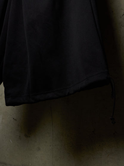Close-up of a black fabric garment, likely a jacket or coat sleeve, hanging against a dimly lit concrete wall with a drawcord detail visible near the bottom right.