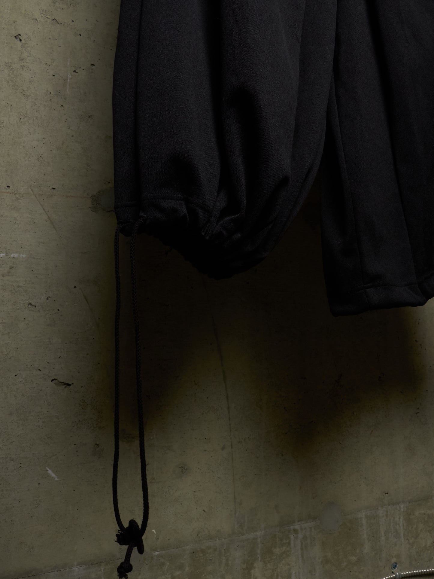 A close-up of black drawstring pants hanging against a textured concrete wall, with the drawstring cord hanging down and a dark shadow cast on the wall.