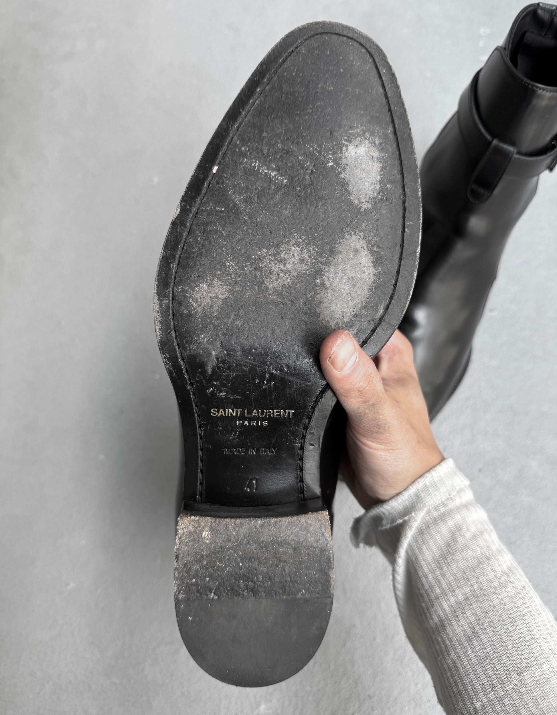 Hand holding a black shoe with 'Saint Laurent' branding on a gray background