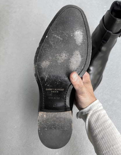 Hand holding a black shoe with 'Saint Laurent' branding on a gray background