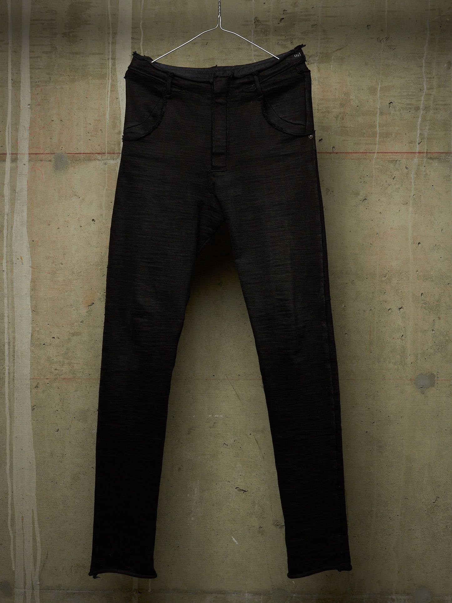 Black slim-fit jeans hanging on a metal hanger against a textured concrete wall.