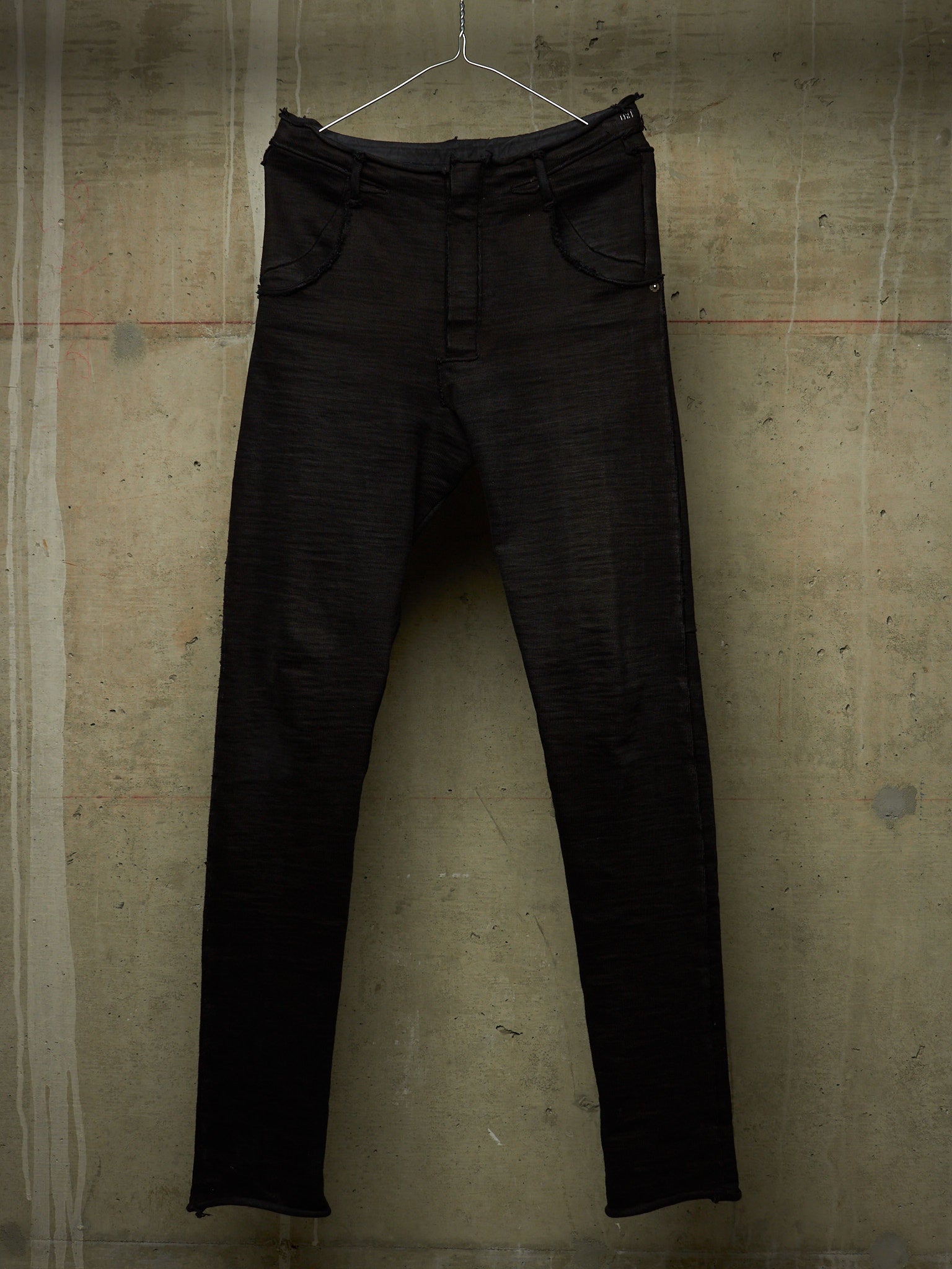 Black slim-fit jeans hanging on a metal hanger against a textured concrete wall.