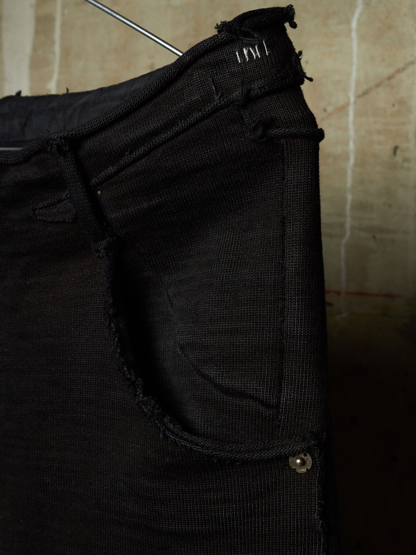Close-up of a black distressed denim garment, likely a skirt or pants leg, hanging against a textured beige wall. The fabric shows heavy fraying, raw edges, and a small metal rivet detail.