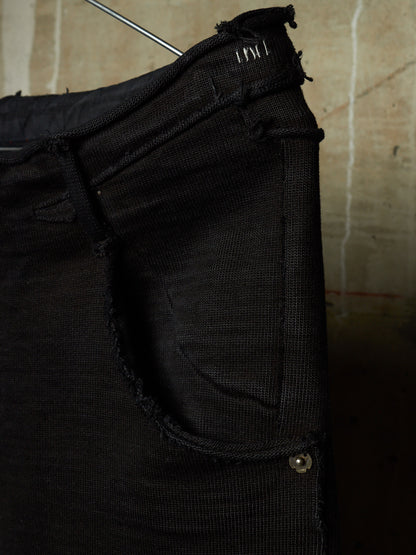 Close-up of a black distressed denim garment, likely a skirt or pants leg, hanging against a textured beige wall. The fabric shows heavy fraying, raw edges, and a small metal rivet detail.