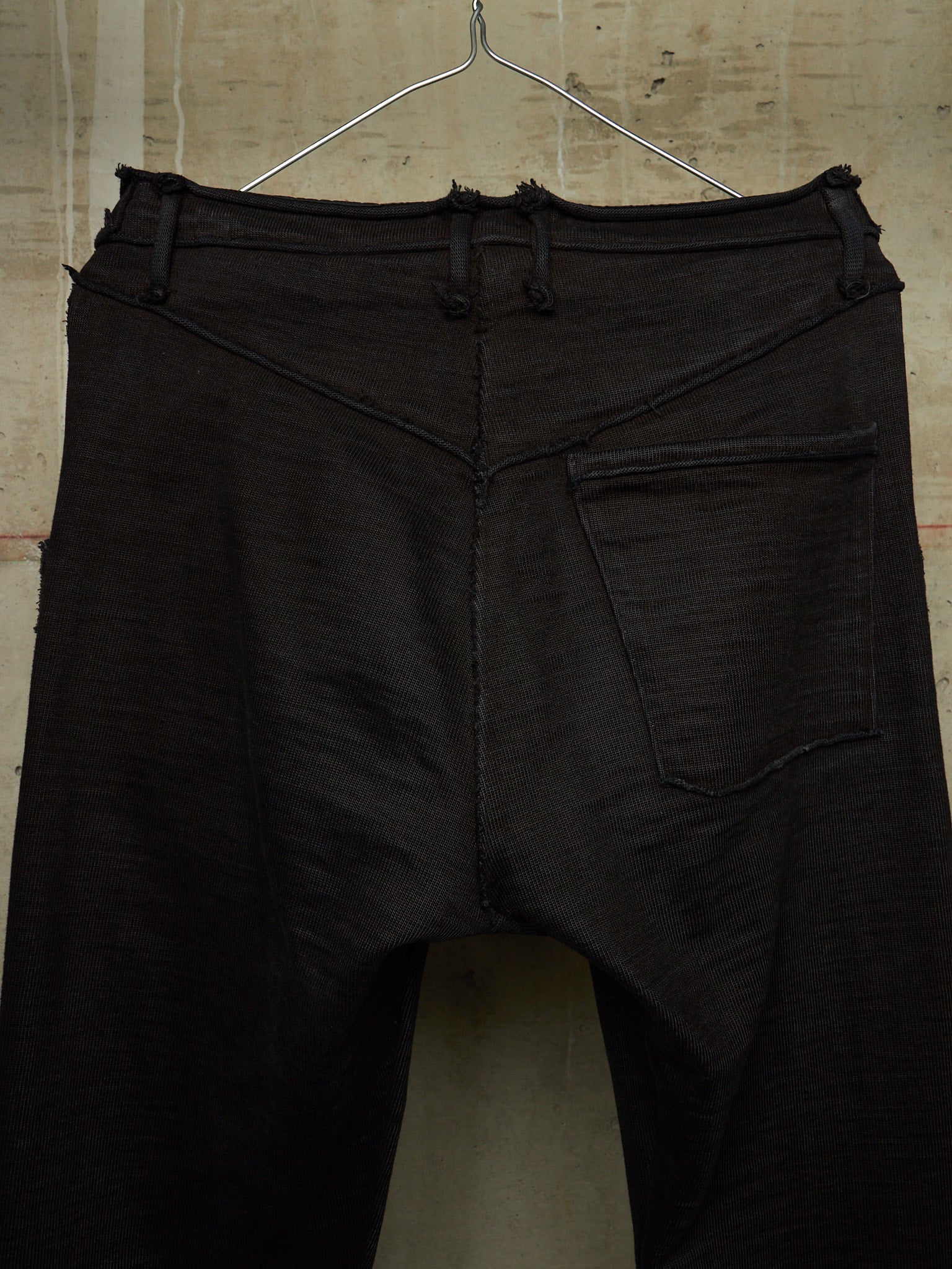 Black distressed denim pants hanging on a wire hanger against a textured concrete wall.