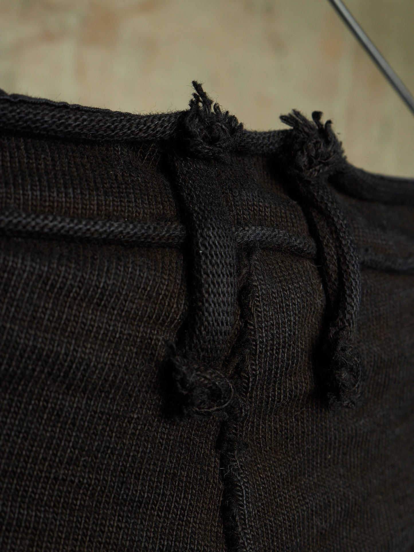 Close-up detail of a dark brown knit garment, showing the waistband, drawstring cords, and frayed tie ends against a softly blurred beige background.
