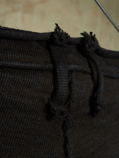 Close-up detail of a dark brown knit garment, showing the waistband, drawstring cords, and frayed tie ends against a softly blurred beige background.
