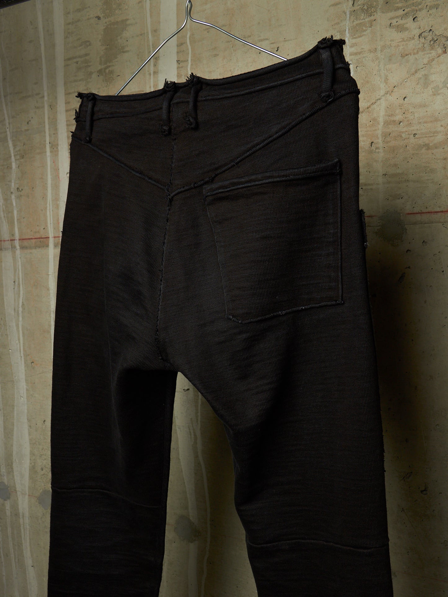 Black textured pants with a unique asymmetrical front seam and a single back patch pocket, hanging on a wire hanger against a textured beige wall.