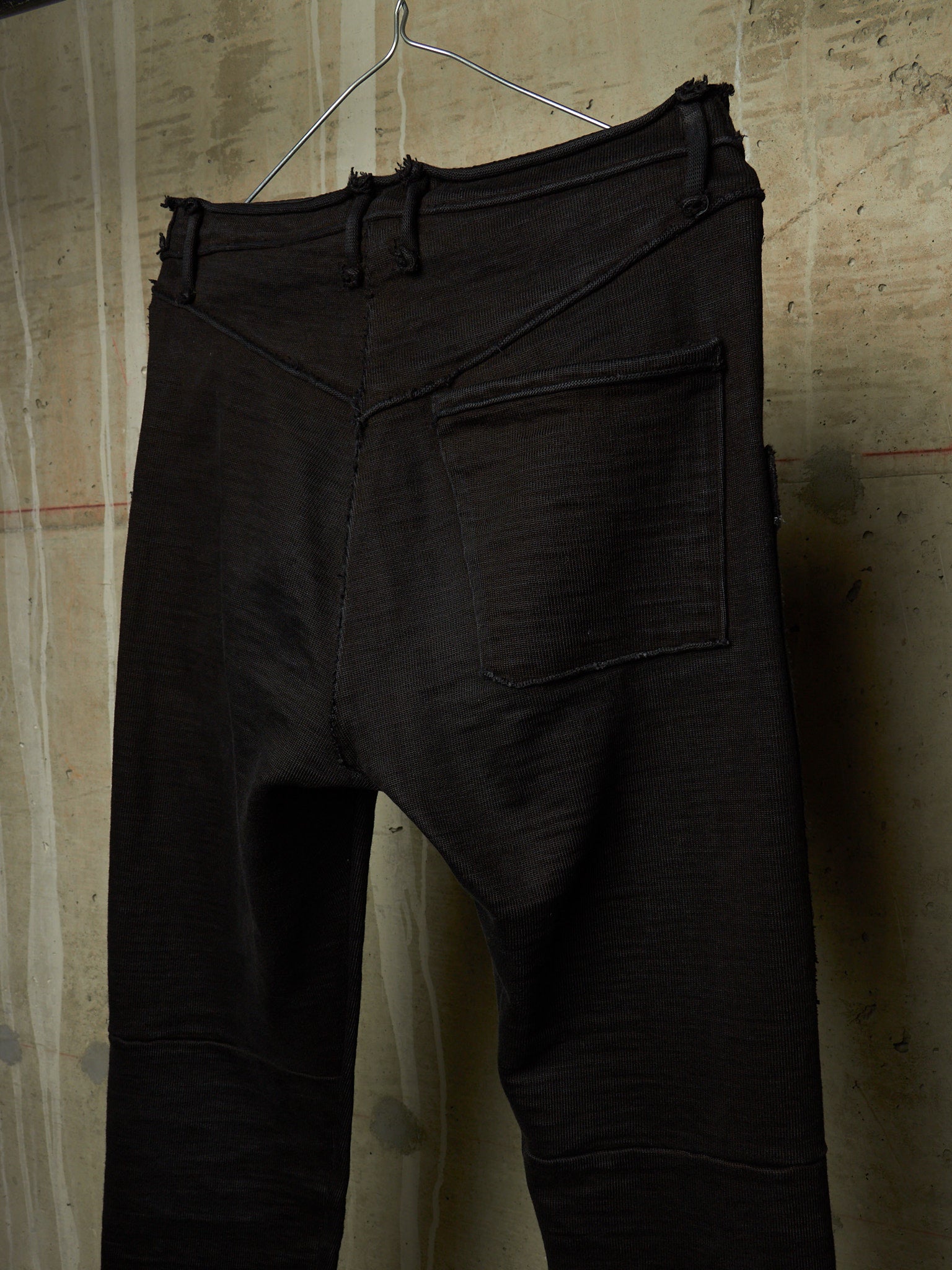 Black textured pants with a unique asymmetrical front seam and a single back patch pocket, hanging on a wire hanger against a textured beige wall.