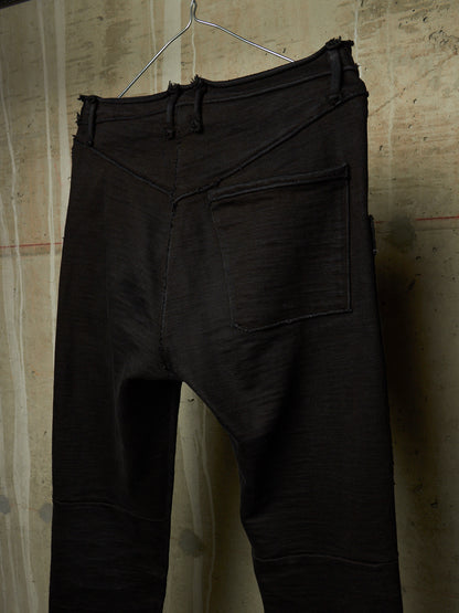 Black textured pants with a unique asymmetrical front seam and a single back patch pocket, hanging on a wire hanger against a textured beige wall.
