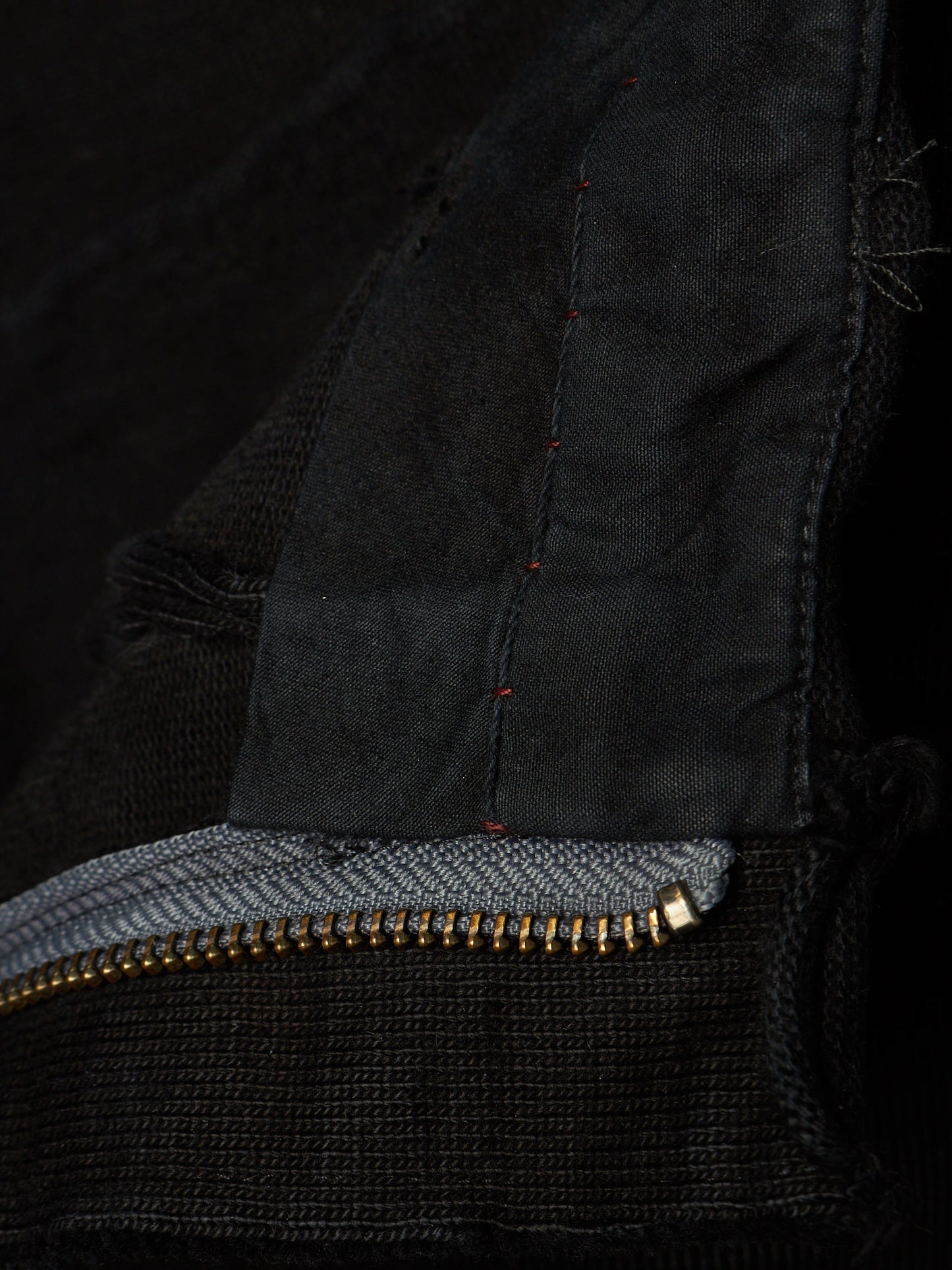 Close-up detail of a black knit garment showing a gold-tone zipper, a gray inner lining, and dark red contrast stitching.