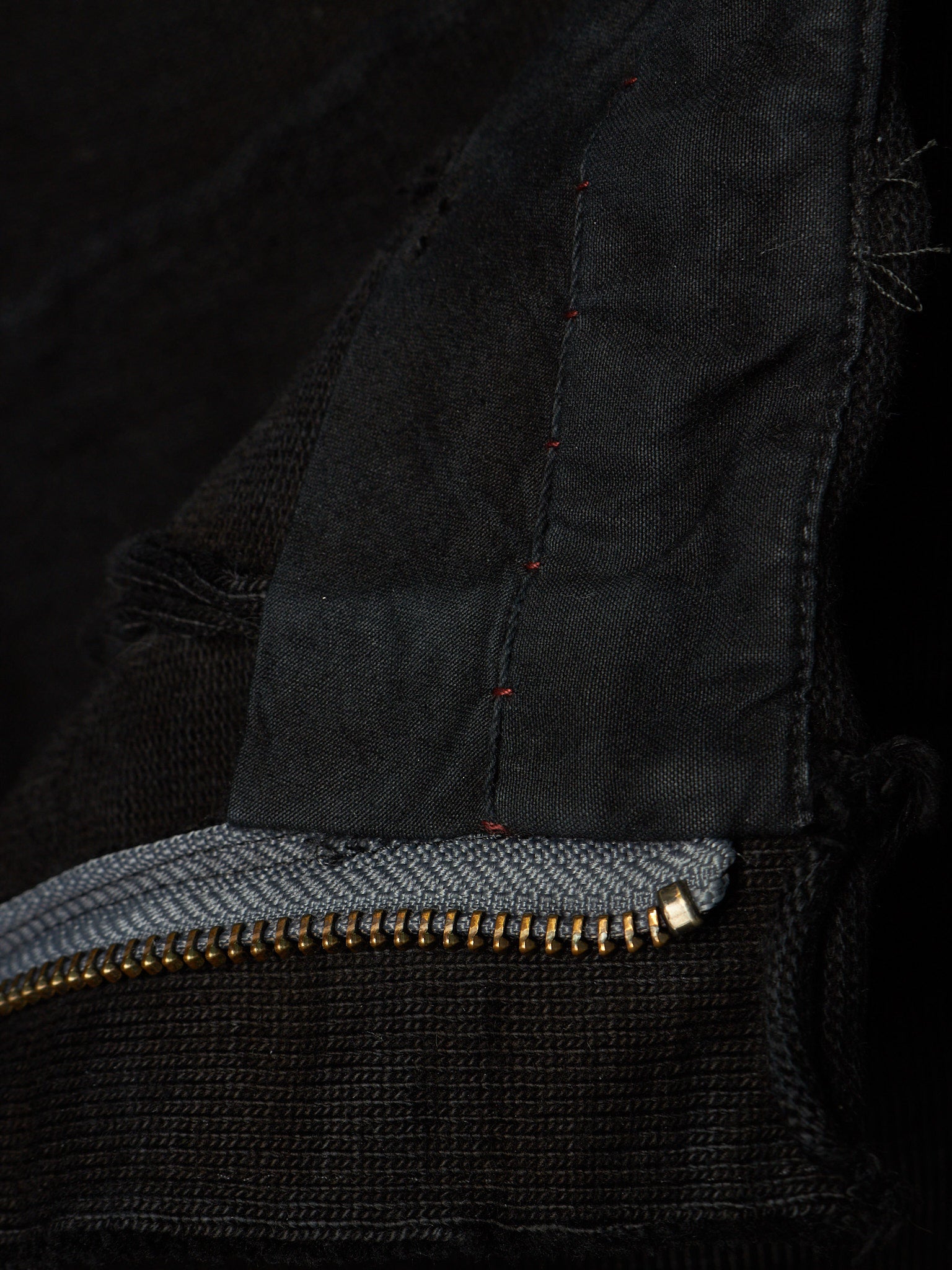 Close-up detail of a black knit garment showing a gold-tone zipper, a gray inner lining, and dark red contrast stitching.