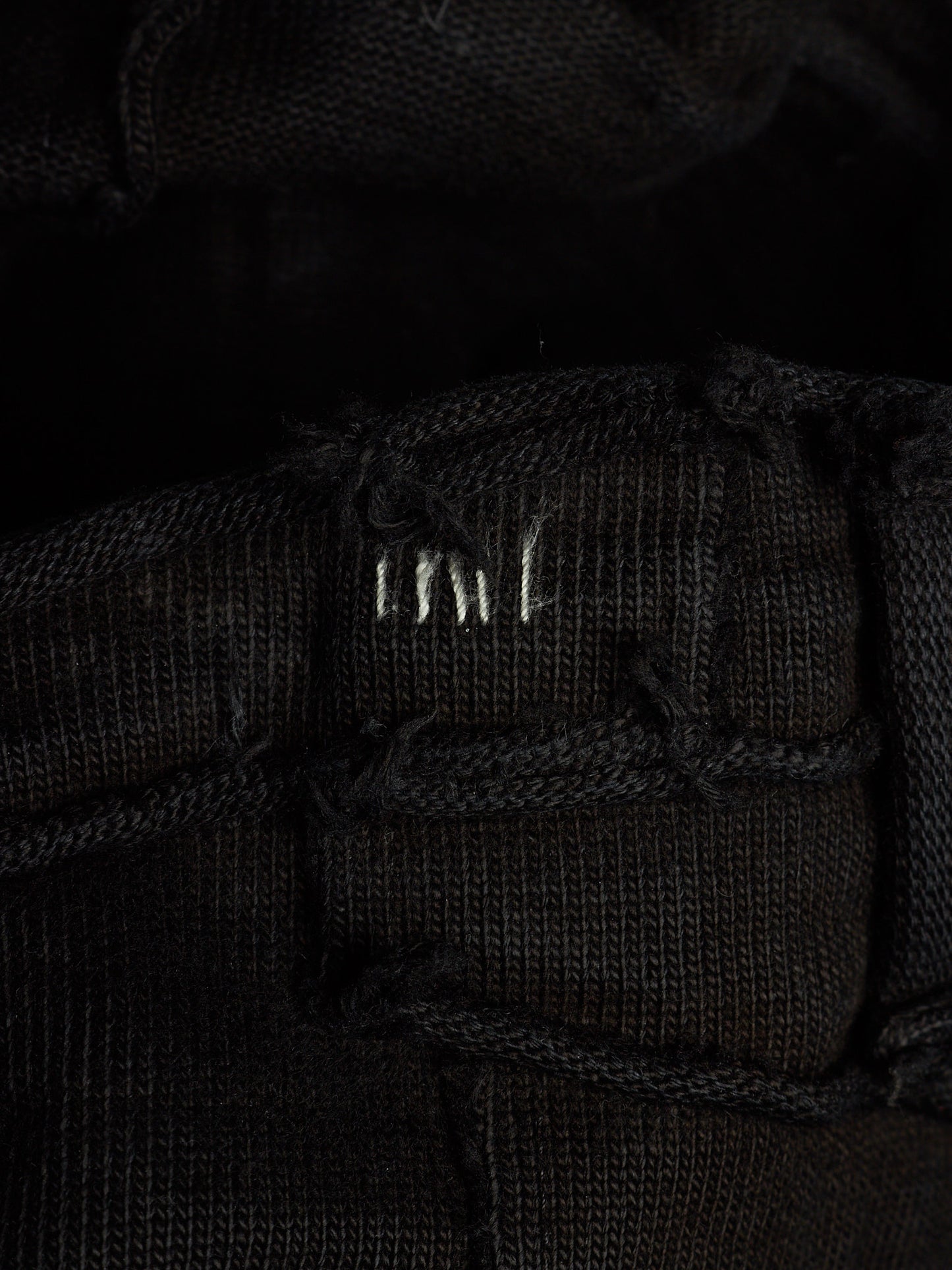 Close-up of a black woven textile with a small white embroidered logo reading 'MNT' on a dark background.