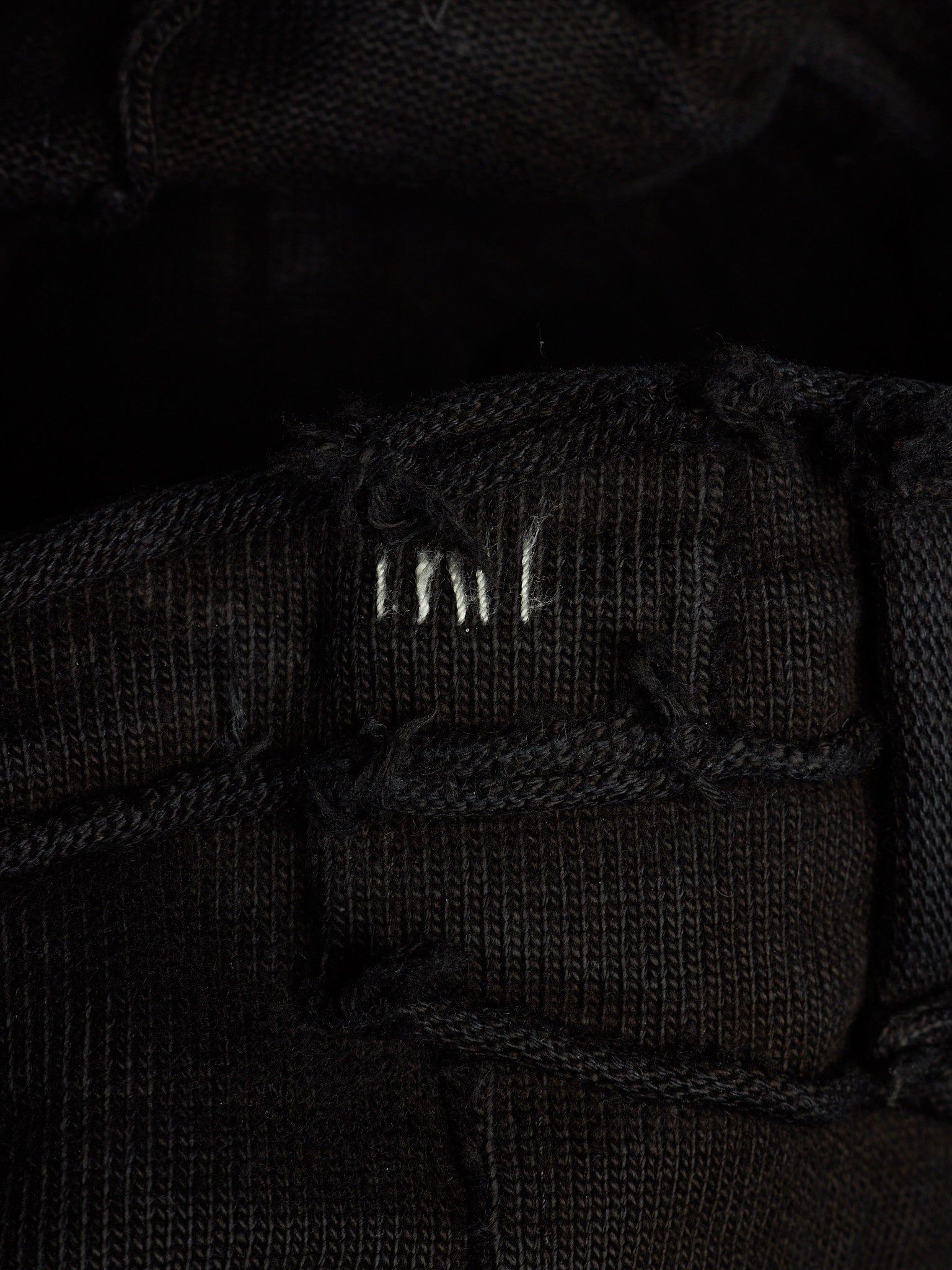 Close-up of a black woven textile with a small white embroidered logo reading 'MNT' on a dark background.