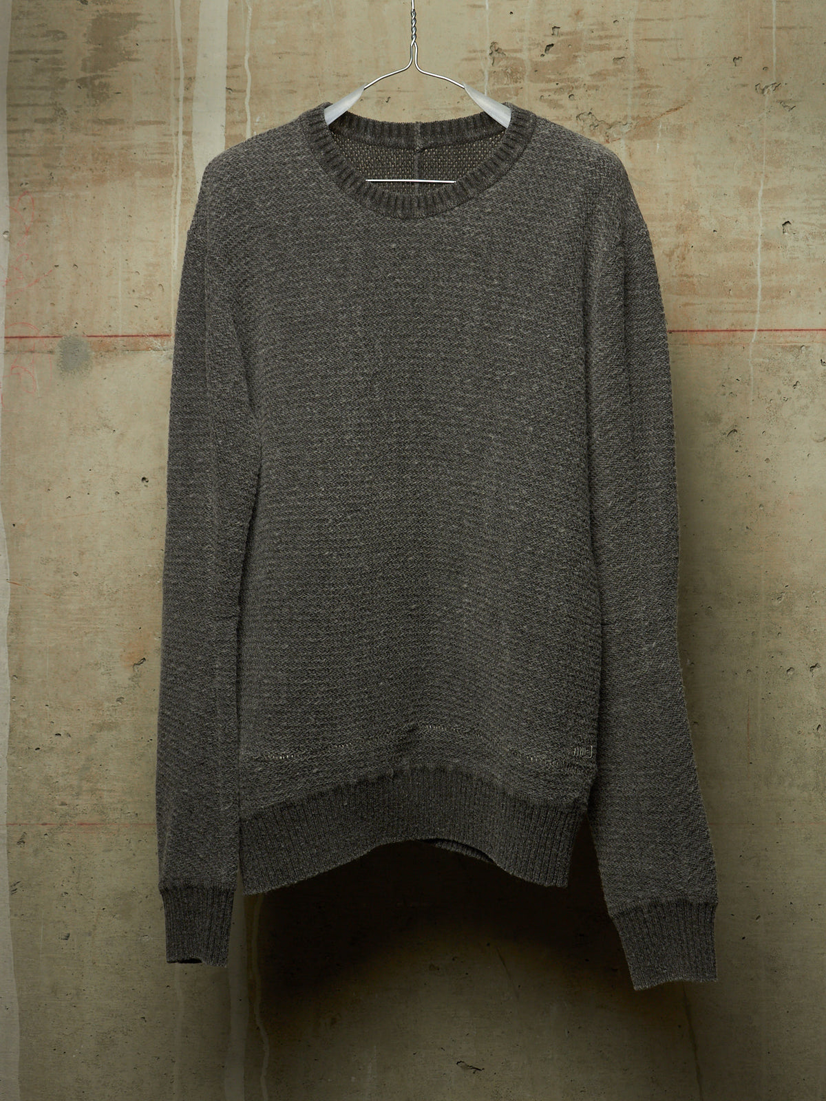 A dark gray textured knit sweater hanging on a white plastic hanger against a rustic concrete wall.