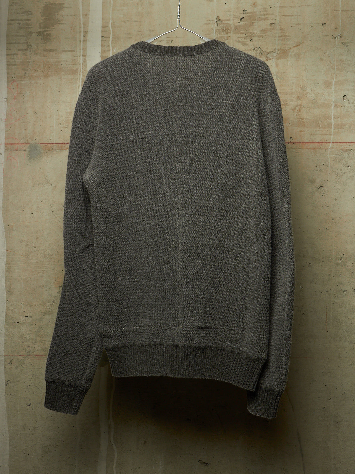 A dark gray textured knit sweater hanging on a metal hanger against a rustic concrete wall.