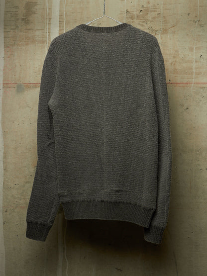 A dark gray textured knit sweater hanging on a metal hanger against a rustic concrete wall.