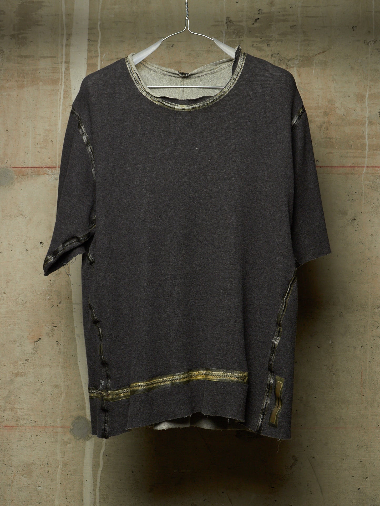 A dark charcoal gray oversized short-sleeve knit top with distressed details, hanging on a white wire hanger against a textured concrete wall.