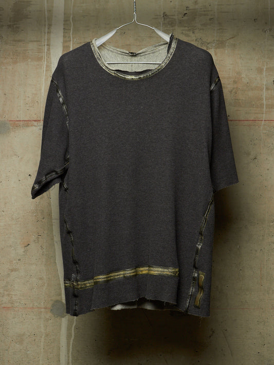 A dark charcoal gray oversized short-sleeve knit top with distressed details, hanging on a white wire hanger against a textured concrete wall.