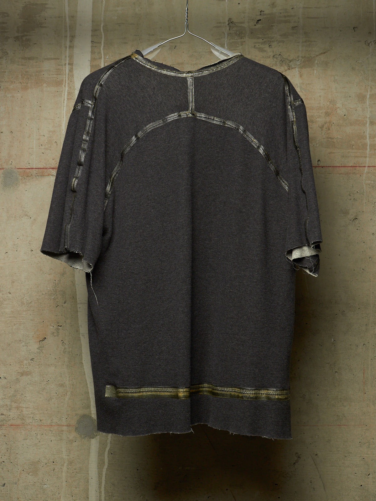 A dark gray oversized short-sleeve top with distressed frayed edges and contrasting light gray patchwork paneling on the back, hanging against a textured concrete wall.