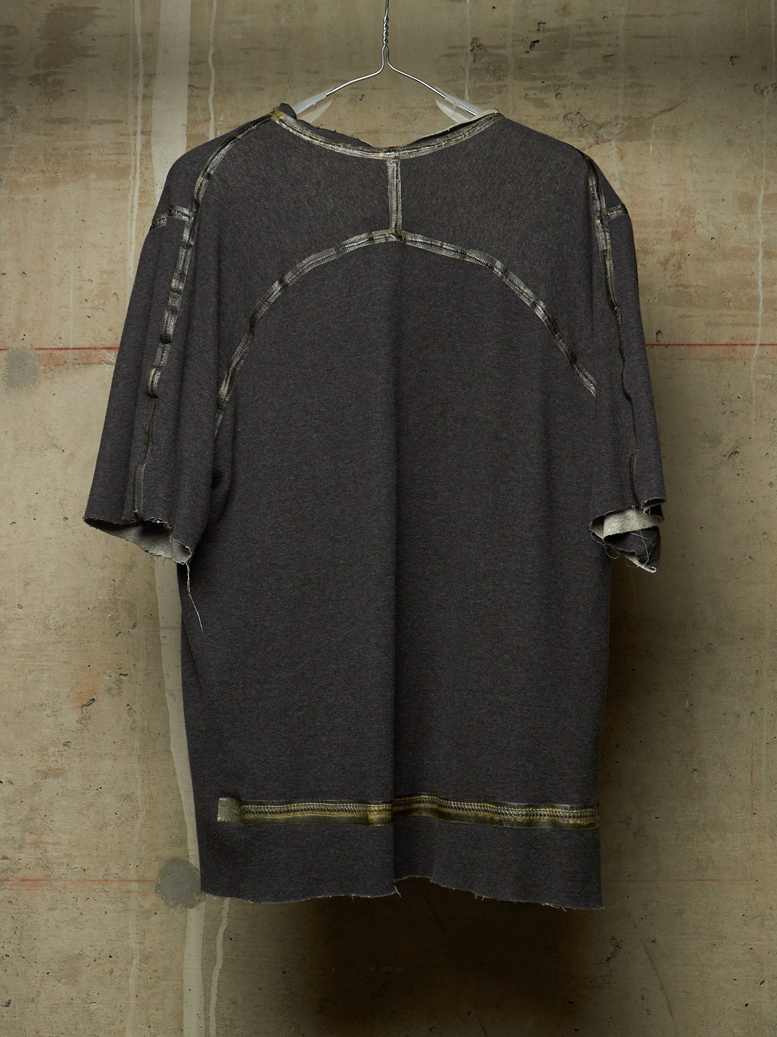 A dark gray oversized short-sleeve top with distressed frayed edges and contrasting light gray patchwork paneling on the back, hanging against a textured concrete wall.