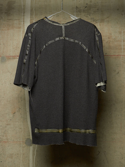 A dark gray oversized short-sleeve top with distressed frayed edges and contrasting light gray patchwork paneling on the back, hanging against a textured concrete wall.