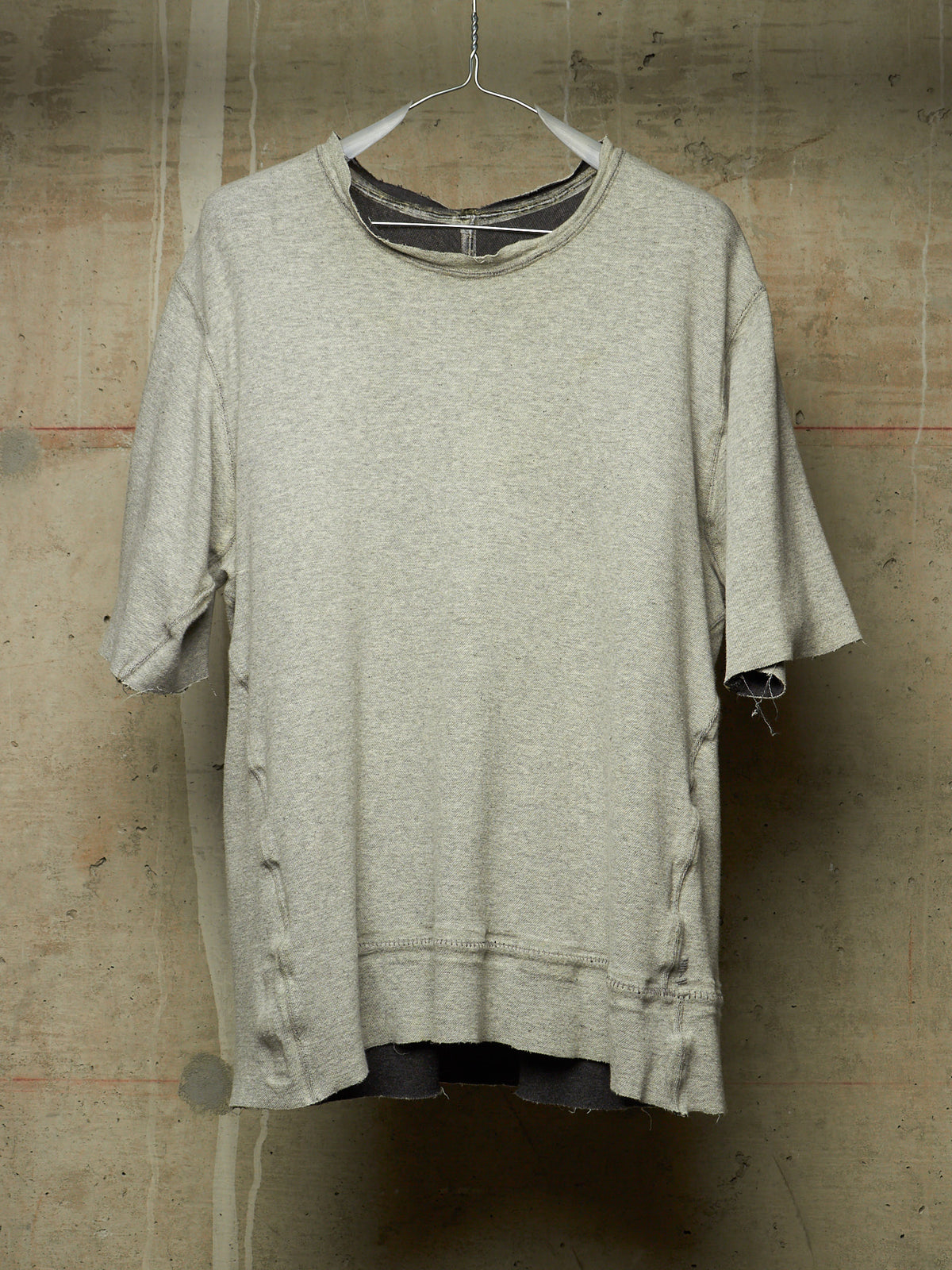 A light gray heather crewneck t-shirt with distressed details, hanging on a wire hanger against a textured concrete wall.