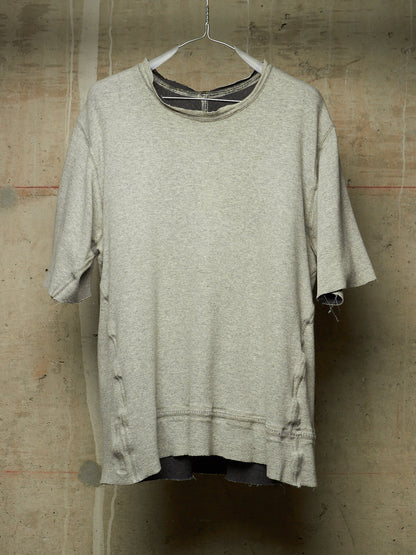 A light gray heather crewneck t-shirt with distressed details, hanging on a wire hanger against a textured concrete wall.