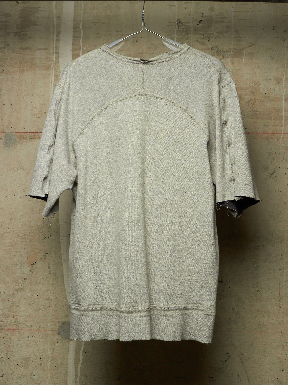 A light gray heathered oversized short-sleeve top with raw, distressed edges and a crew neck, hanging on a wire hanger against a textured concrete wall.
