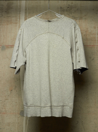 A light gray heathered oversized short-sleeve top with raw, distressed edges and a crew neck, hanging on a wire hanger against a textured concrete wall.