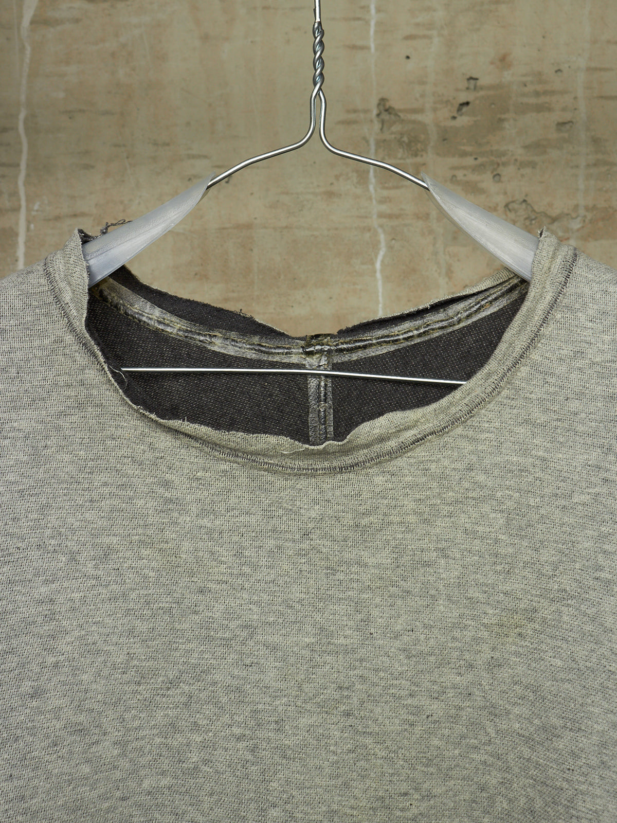 A close-up of a heather gray crewneck sweatshirt hanging on a white plastic hanger against a textured beige wall.