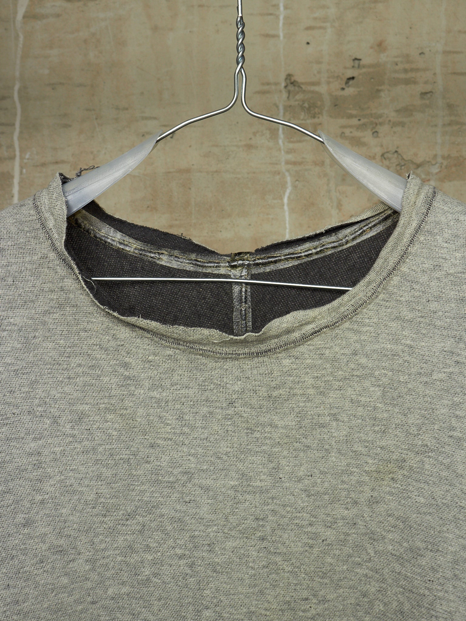 A close-up of a heather gray crewneck sweatshirt hanging on a white plastic hanger against a textured beige wall.
