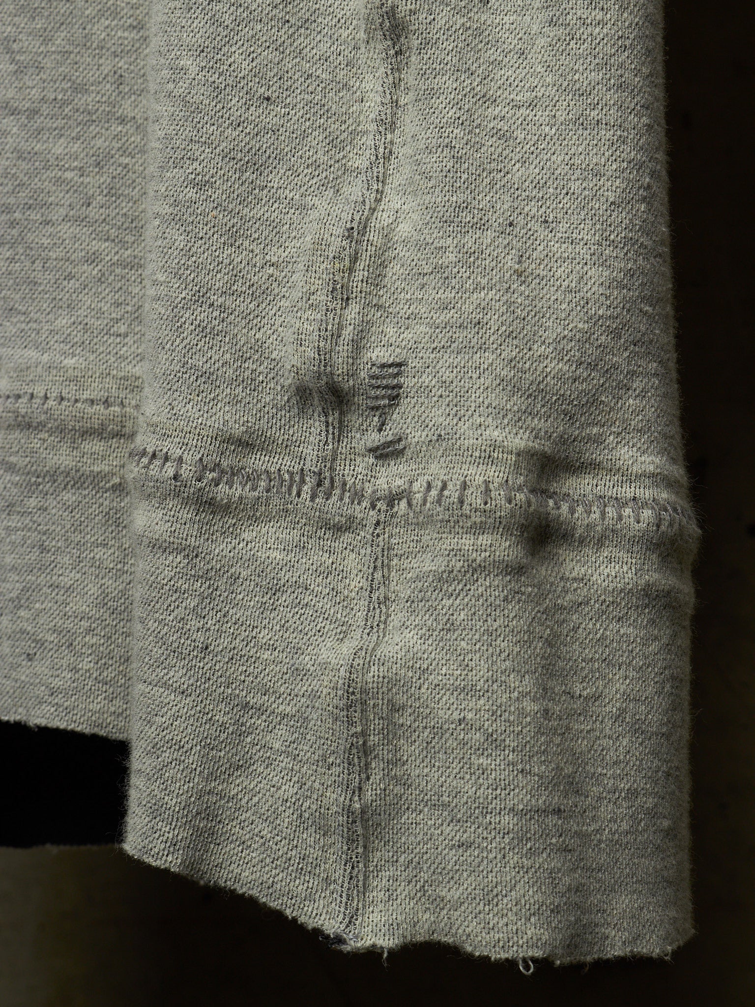 Close-up of a light gray knit fabric sleeve or cuff with visible stitching and a small dark embroidered detail near the center.
