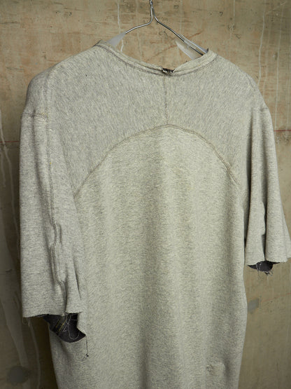 A light gray heathered oversized knit top hanging on a metal hanger against a textured concrete wall. The shirt features a crew neck, a curved seam detail across the upper chest, and raw, frayed edges on the sleeves.