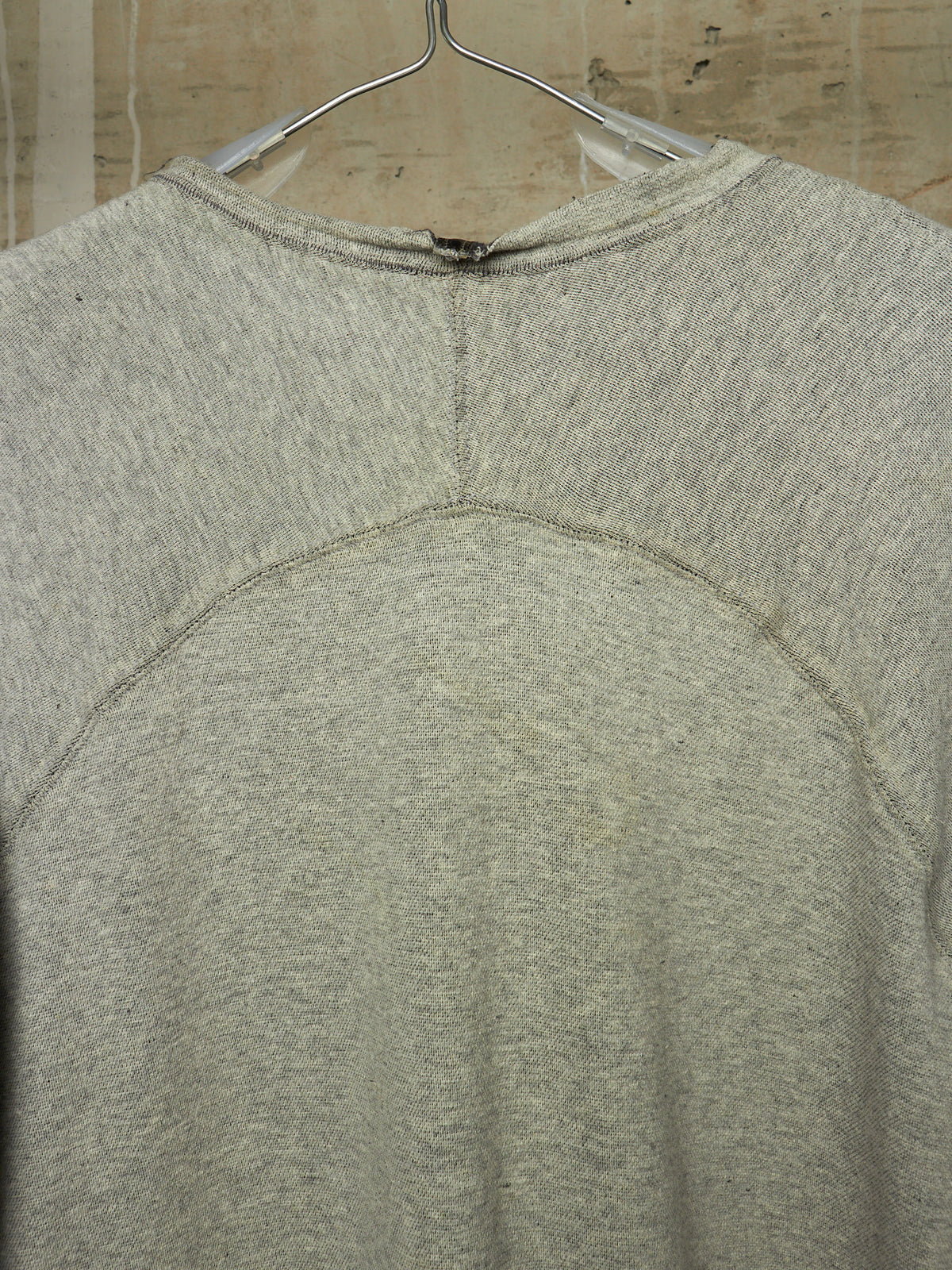 A close-up of the back of a light gray heather crewneck sweatshirt hanging on a metal hanger against a beige tiled wall.