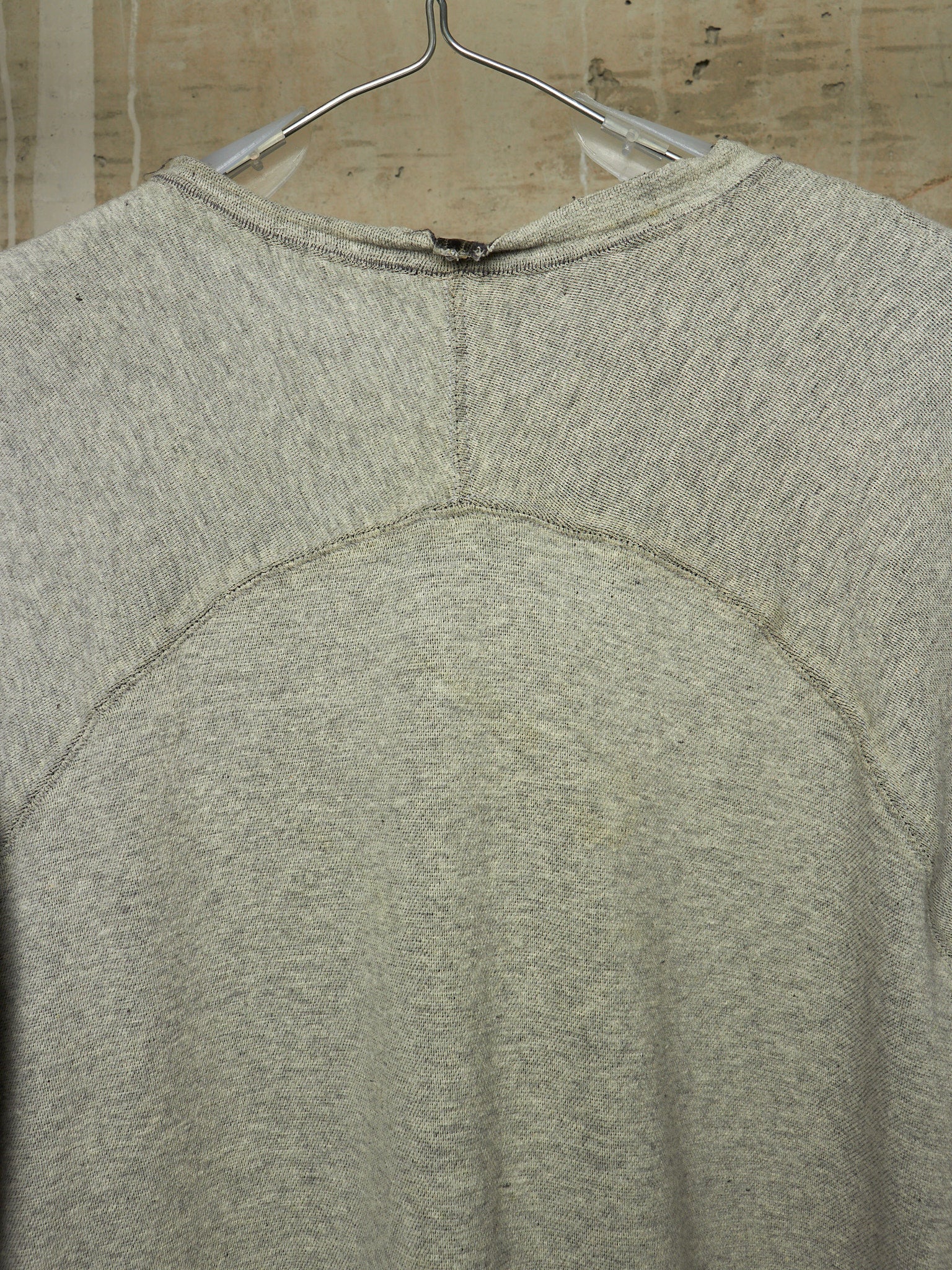 A close-up of the back of a light gray heather crewneck sweatshirt hanging on a metal hanger against a beige tiled wall.