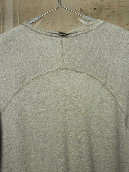A close-up of the back of a light gray heather crewneck sweatshirt hanging on a metal hanger against a beige tiled wall.