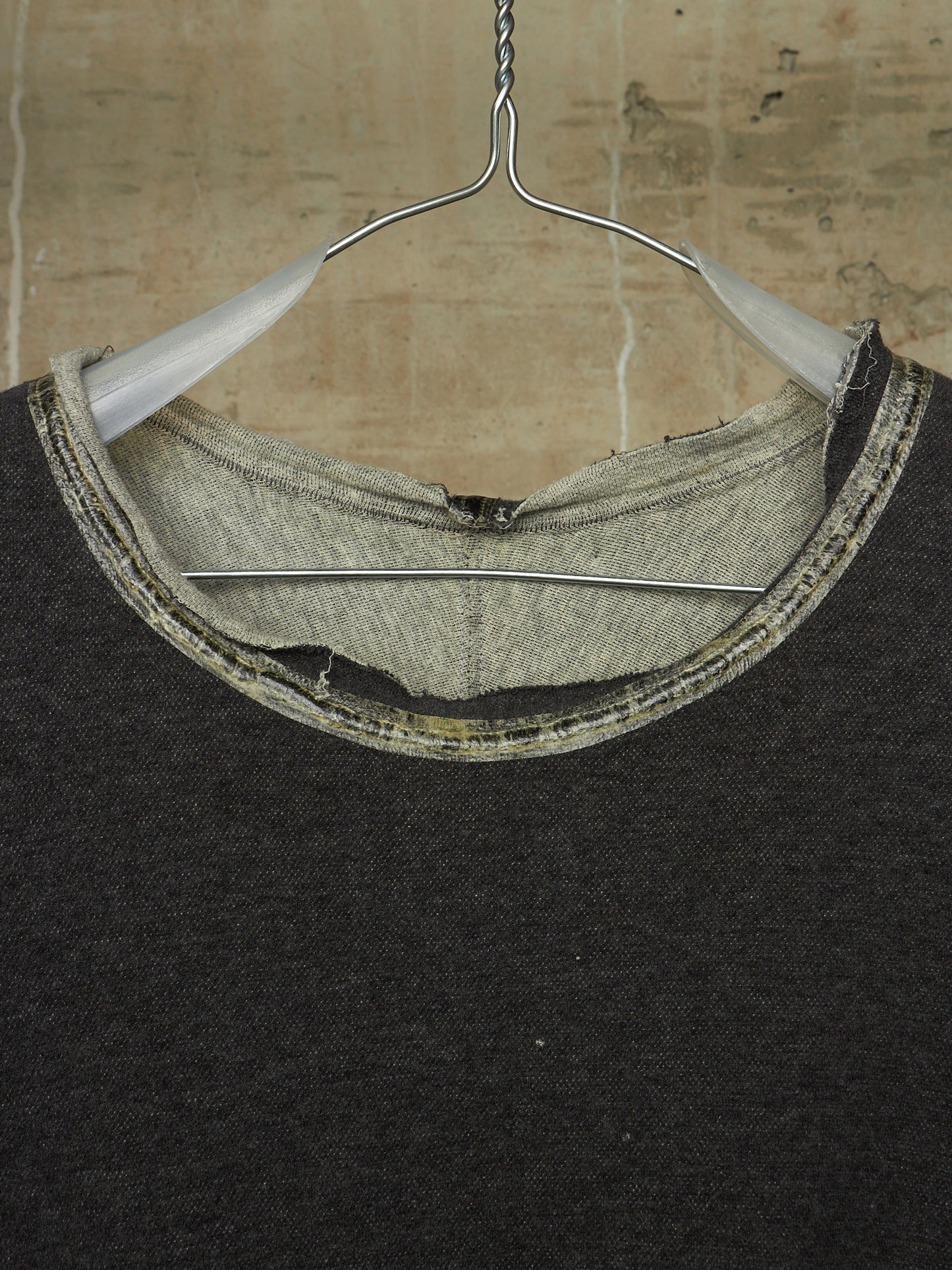 Close-up of a dark charcoal knit crewneck sweater hanging on a metal hanger against a textured beige wall. The neckline shows a raw, unfinished hem with a contrasting light gray inner lining and a stitched trim.