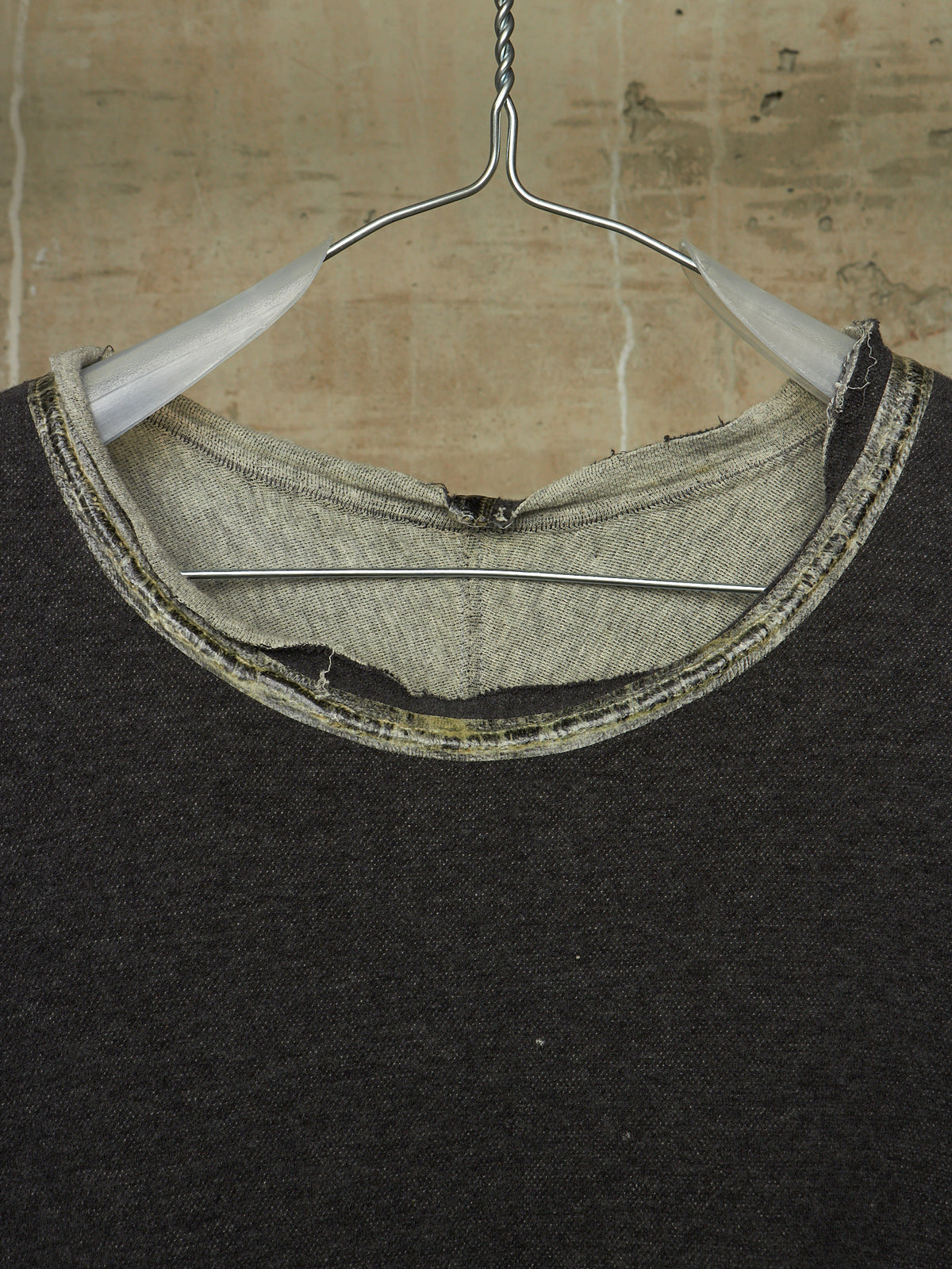 Close-up of a dark charcoal knit crewneck sweater hanging on a metal hanger against a textured beige wall. The neckline shows a raw, unfinished hem with a contrasting light gray inner lining and a stitched trim.
